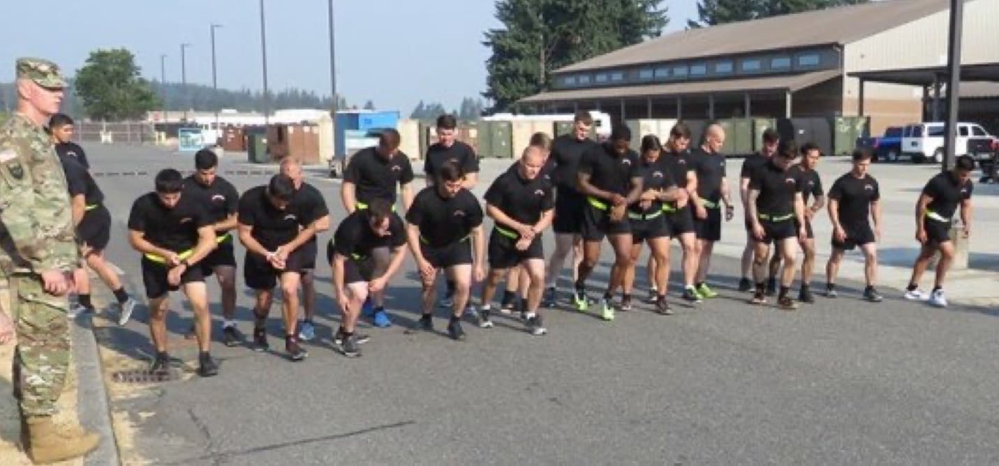 A line of Soldiers in black PT uniforms crouch at a start line on pavement, ready to run, while a grader in uniform stands at the side.