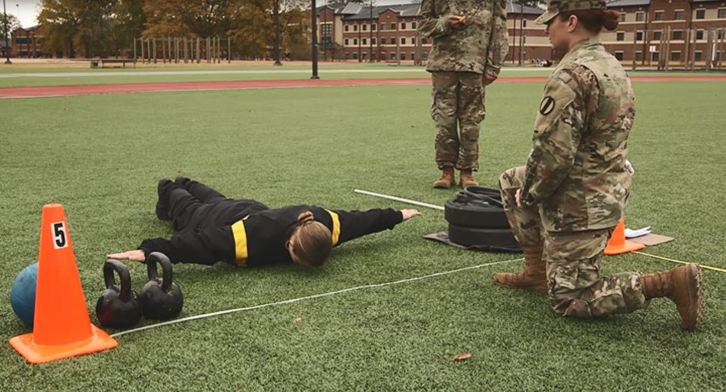 A Soldier lies prone on turf with both arms extended straight out to the sides in the hand-release position while a grader kneels beside the lane watching; another grader stands behind.