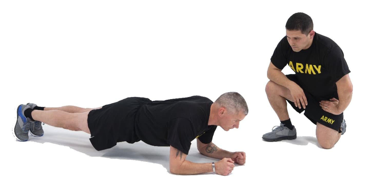 A Soldier holds a forearm plank with fists on the ground, elbows under the shoulders, and the body straight from head to heels on the toes, while a grader kneels nearby watching.